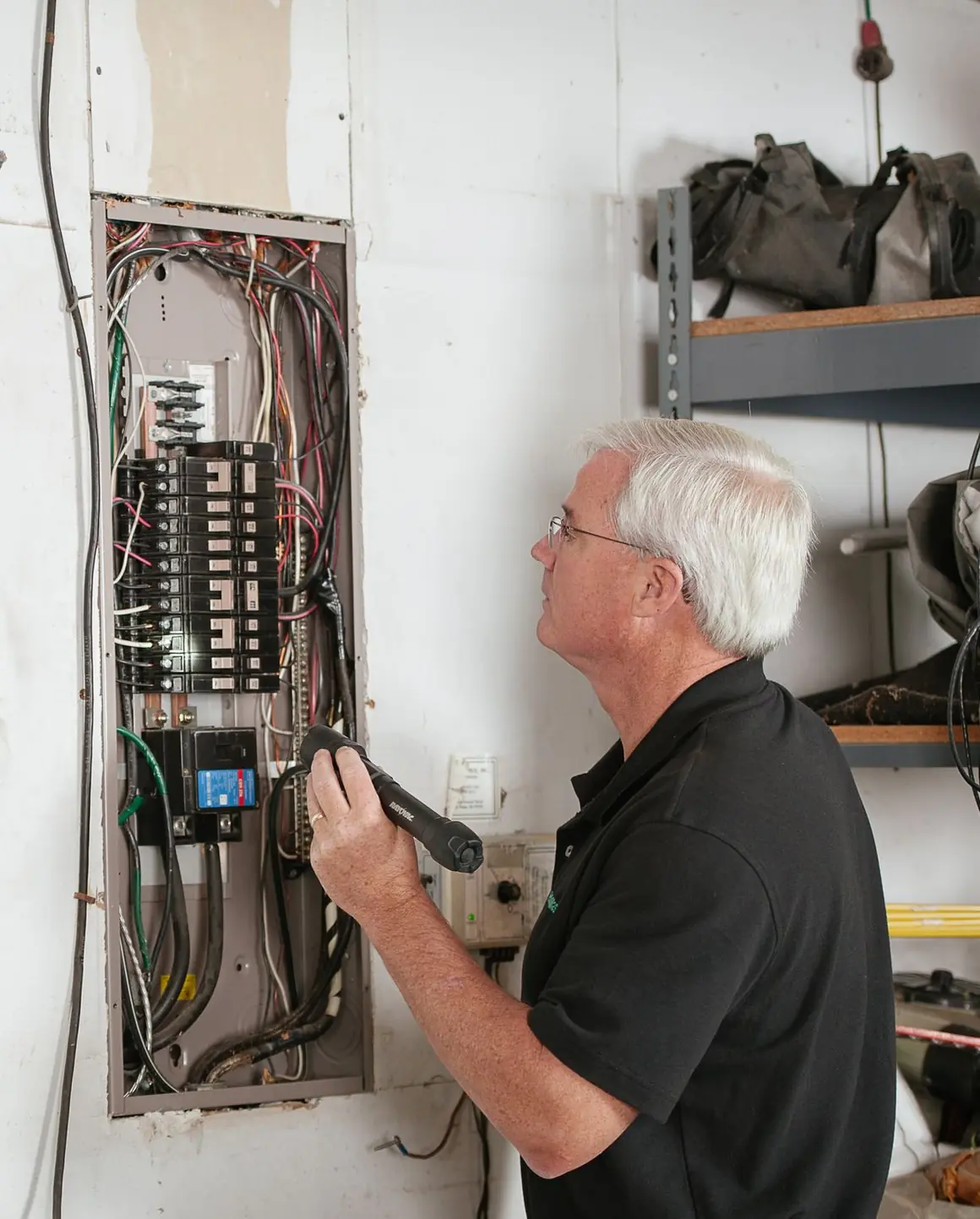 An experienced electrician in a black polo shirt inspecting a residential switchboard with a test instrument.